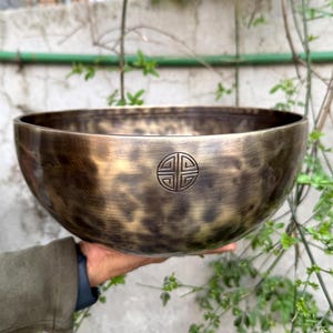 May include: A large, round, antique-looking singing bowl with a hammered finish. The bowl is made of metal and has a dark brown color with a gold sheen. The bowl has a symbol engraved on the bottom.