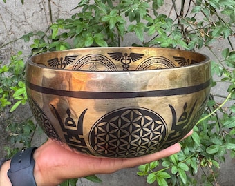 May include: A large, golden singing bowl with intricate black designs. The bowl features a flower of life pattern, a caduceus symbol, and a geometric pattern. The bowl is being held in a hand.