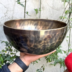 May include: A large, round, antique-looking singing bowl with a hammered finish. The bowl is made of metal and has a dark brown color with a hint of gold. The bowl is being held by a hand.