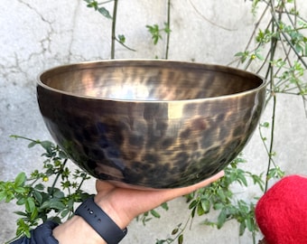 May include: A large, round, antique-looking singing bowl with a hammered finish. The bowl is made of metal and has a dark brown color with a hint of gold. The bowl is being held by a hand.