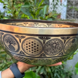 Może przedstawiać: Misa do śpiewania w kolorze mosiądzu ze skomplikowanymi czarnymi wzorami. Misa posiada różne okrągłe wzory, w tym drzewo życia, kształty geometryczne i smoka. Obręcz ma ciemnobrązowy pasek.