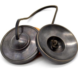 May include: A pair of antique-looking cymbals with a leather strap connecting them. The cymbals are dark gray and have a slightly textured surface.