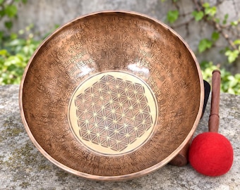 May include: A round bronze singing bowl featuring an etched flower of life geometric pattern at the center and intricate Sanskrit mantra inscriptions covering the interior walls, accompanied by a wooden mallet with a red felt-covered head.