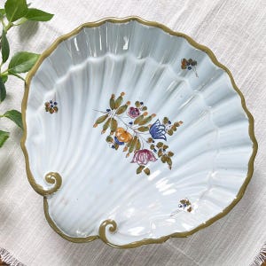 May include: A white, shell-shaped decorative plate with a scalloped edge and a gold trim. The plate features a hand-painted floral design in shades of blue, orange, and purple. The plate is a decorative item.