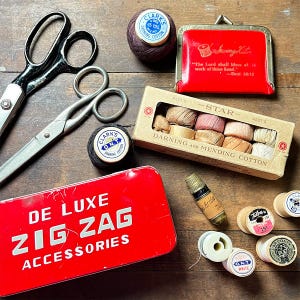 May include: A collection of vintage sewing supplies including two pairs of metal scissors, a red metal De Luxe Zig Zag Accessories tin, a red coin purse with text, a box of Star darning and mending cotton, a thimble, and various spools of thread.