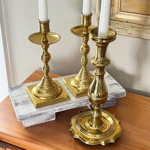May include: Three ornate brass candlesticks with white candles. The candlesticks have a polished finish and decorative detailing. They are arranged on a white wooden tray, creating a classic and elegant display. The candlesticks vary in height.