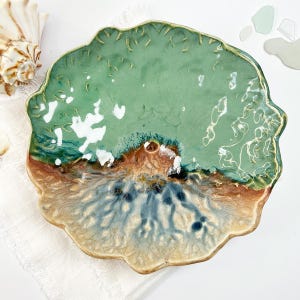May include: A handcrafted ceramic plate with a unique, organic shape. The plate features a blend of colors, including seafoam green, brown, and blue, with a glossy finish. The edges have a textured, scalloped design, adding to its artistic appeal. A decorative piece for the home.