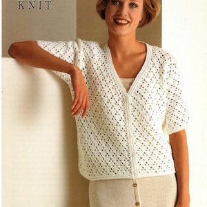 Könnte beinhalten: Ein weißer Häkelcardigan mit V-Ausschnitt und kurzen Ärmeln. Der Cardigan ist mit einer Doppelstrickmasche gearbeitet und hat einen Knopfverschluss. Das Model trägt den Cardigan mit einem weißen Tanktop und einem weißen Rock.