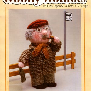 May include: Knitting pattern for a knitted doll named Grandad. The doll is wearing a brown coat, a red hat, and a scarf. The doll is holding a pipe in its mouth and is standing at a bus stop. The pattern is number 026 and the doll is approximately 30 cm tall.