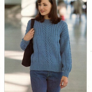 May include: A blue knitted jumper with a cable knit pattern. The jumper has a round neckline and long sleeves. The model is wearing blue jeans.