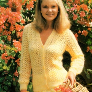 May include: A woman wearing a yellow knitted cardigan with a V-neck and button closure. The cardigan has a textured pattern. She is holding a woven basket with flowers. The background features orange flowers and greenery. The text "Gemini/DK" and "32-44" are visible.