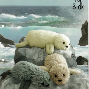 May include: Three plush toy seals on a rock with a blue ocean and sky in the background. The seals are white, brown, and gray. The text "Luxe Fur & dk" is in the upper right corner of the image.