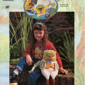 Op de afbeelding: Een meisje draagt een rode trui met een gebreid kikkerontwerp en zit met een gebreide kikkerpop. De pop draagt een bruin vest en een geruit overhemd. Het meisje houdt de hand van de pop vast. De achtergrond is een groene graszone met een bakstenen muur.