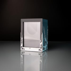 Op de afbeelding: Een vierkante, grijze en blauwgroene fotolijst met een heldere glazen voorkant. De lijst heeft een donkergrijze bovenkant en een blauwgroene zijkant met een textuur, bubbelachtig patroon. De lijst staat op een reflecterend oppervlak.