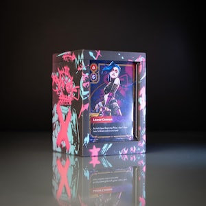 Op de afbeelding: Een kaarten display box met een karakterkaart zichtbaar door een venster. De kaart heeft een karakterillustratie en de tekst "Loose Cannon". De doos heeft een zwarte achtergrond met roze en turquoise graffiti-achtige ontwerpen.