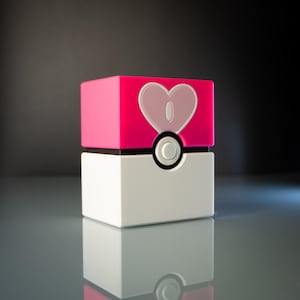 Könnte beinhalten: Ein rosa und weißer Pokéball mit einem Herzdesign auf der Oberseite. Der Pokéball ist ein beliebtes Sammlerstück für Fans des Pokémon-Franchise.
