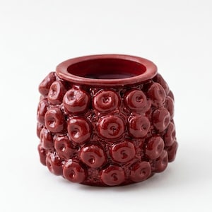 Può includere: Un piccolo vaso rotondo in ceramica bordeaux con una superficie strutturata. Il vaso è ricoperto da piccoli disegni circolari in rilievo e ha una parte superiore liscia e aperta. Il vaso è probabilmente utilizzato per scopi decorativi.