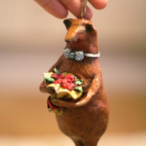 Può includere: Figurina marrone a forma di orso che tiene in mano un bouquet di rose rosse. L'orso indossa un papillon a pois blu e bianchi e un colletto bianco. L'ornamento è sospeso da uno spago marrone, probabilmente per decorare.