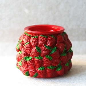 May include: A red ceramic container with a wide opening and a base covered in 3D strawberry designs. The strawberries are a vibrant red with green leaves, creating a textured, fruity appearance. The container is likely used for decorative purposes.