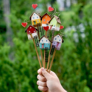 Op de afbeelding: Een boeket van kleurrijke miniatuurhuisjes op houten stokjes, elk met een kleine ladder en een hartvormig accent. De huisjes zijn geschilderd in verschillende kleuren, waaronder geel, oranje, groen en rood, en worden in een hand gehouden tegen een vage groene achtergrond.