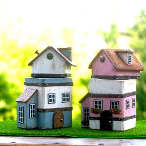 Könnte beinhalten: Zwei dekorative Miniaturhäuser. Eines ist weiß und blau, das andere rosa und weiß. Beide haben mehrere Stockwerke, Fenster und dekorative Dächer. Sie stehen auf einem Stück grünen Grases.