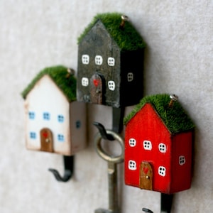Peut inclure: Trois maisons miniatures en bois avec des toits en mousse verte et des crochets en métal. Chaque maison est peinte d'une couleur différente : noir, blanc et rouge. Les maisons ont de petites fenêtres blanches et un cœur rouge sur la porte. Une clé est accrochée aux crochets.