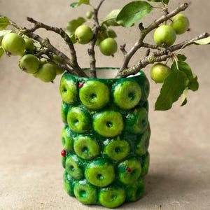 Peut inclure: Un vase cylindrique en céramique verte avec une surface texturée recouverte de pommes vertes moulées et de petites coccinelles rouges. Plusieurs branches avec des feuilles et des petites pommes vertes sortent du vase, une pomme est posée à côté.