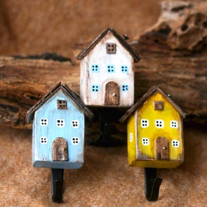 Puede incluir: Tres casas de madera en miniatura pintadas en azul, blanco y amarillo con detalles negros. Cada casa tiene una pequeña puerta y ventanas. Las casas están unidas a ganchos de metal negro.