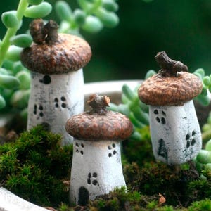 Op de afbeelding: Drie miniatuurhuisjes met wit getextureerde lichamen en daken van bruine eikeldopjes. Elk huisje is voorzien van kleine, met de hand geschilderde zwarte ramen en een deur, met een piepklein takje dat uit het midden van de eikeldop steekt.