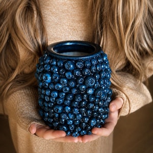 Può includere: Un vaso cilindrico ricoperto da uno strato denso e strutturato di mirtilli blu scuro realistici. La superficie presenta numerose bacche individuali di varie dimensioni, creando un aspetto organico e irregolare con un bordo superiore liscio.