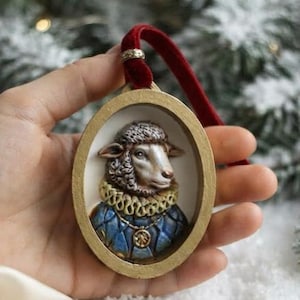 Puede incluir: Un adorno de forma ovalada con un relieve tridimensional de una oveja que lleva un cuello con volantes y una prenda azul acolchada. La oveja está enmarcada por un borde dorado y cuelga de una cinta de terciopelo rojo con un anillo plateado.