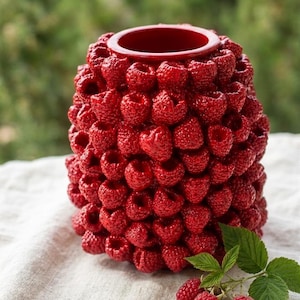 May include: A red ceramic vase with a textured surface, designed to resemble a cluster of raspberries. The vase has a cylindrical shape with a wide opening at the top. A few fresh raspberries and green leaves are placed near the vase.