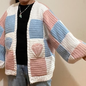 May include: A white, pink, and blue patchwork cardigan with a diamond-shaped design on the front pockets. The cardigan has a loose fit and long sleeves.