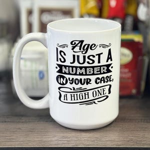 May include: White ceramic coffee mug with black text that reads "Age is just a number. In your case, a high one."