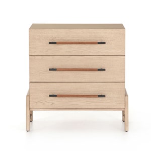 Könnte beinhalten: Eine hellfarbene Holzkommode mit drei Schubladen. Jede Schublade hat einen braunen, lederummantelten Griff mit schwarzen Metallakzenten. Die Kommode steht auf kurzen Beinen.