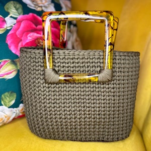 May include: A green crocheted tote bag with a tortoise shell plastic handle. The bag is sitting on a yellow couch.