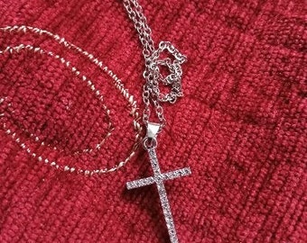 May include: A silver chain necklace with a silver cross pendant encrusted with small clear stones. The necklace is lying on a red fabric background.