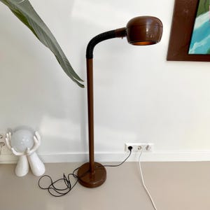 May include: A brown mid-century modern floor lamp with a round base and a curved, adjustable neck. The lamp has a cylindrical stem and a rounded lampshade. The lamp is plugged into a wall outlet. A white decorative object is on the floor.