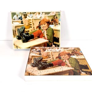 May include: Two puzzles featuring a scene of a girl with red braids in a classroom setting. The girl is wearing a green jacket, orange socks, and black boots. The puzzles are in white boxes.