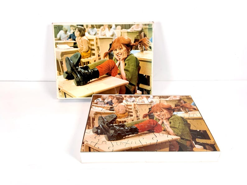 May include: Two puzzles featuring a scene of a girl with red braids in a classroom setting. The girl is wearing a green jacket, orange socks, and black boots. The puzzles are in white boxes.