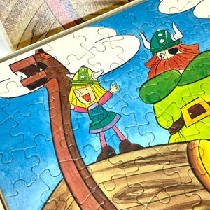 Könnte beinhalten: Ein farbenfrohes Puzzle mit einem Cartoon-Wikinger mit Augenklappe und einem Mädchen mit blonden Haaren. Der Wikinger trägt ein grünes und braunes Kostüm und hält eine Axt. Das Mädchen trägt ein grünes und gelbes Kleid und hält ein Schwert. Die Puzzleteile sind in Form eines Schiffsrumpfes angeordnet.
