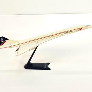 Può includere: Modello di aereo Concorde color crema con dettagli rossi e neri e il logo British Airways. Il modello è montato su un supporto nero con la scritta "Concorde" sulla base. Le ali dell'aereo sono rivolte verso l'alto.