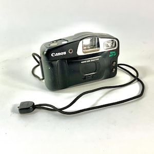 May include: A black Canon Prima BF-7 film camera with a black strap. The camera has a silver lens and a green label that says "Prima BF-7".