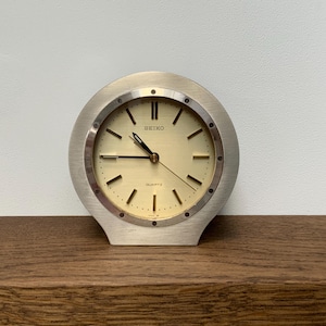 May include: A silver Seiko quartz clock with a gold face. The clock features black hands and hour markers. It is displayed on a wooden surface.
