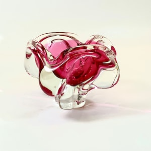 Op de afbeelding: Een heldere glazen schaal met een levendig roze interieur. De schaal heeft een unieke, organische vorm met wervelende, bloembladachtige randen. Het glas is dik en lijkt mondgeblazen, wat een mix van transparantie en kleur laat zien.