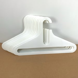 Puede incluir: Un conjunto de perchas de plástico blanco. Las perchas tienen un diseño curvo con una zona ancha para los hombros y un gancho para colgar la ropa. Las perchas son de material liso y sólido, apiladas ordenadamente.