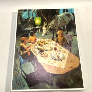May include: A rectangular jigsaw puzzle box featuring a vintage illustration of two children sitting at a wooden dining table with a white horse standing beside them. A green glass pendant light hangs above the table, which is set with food.