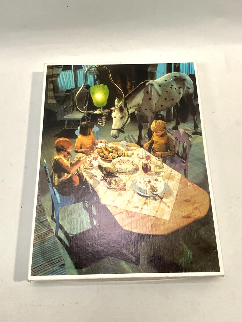 May include: A rectangular jigsaw puzzle box featuring a vintage illustration of two children sitting at a wooden dining table with a white horse standing beside them. A green glass pendant light hangs above the table, which is set with food.