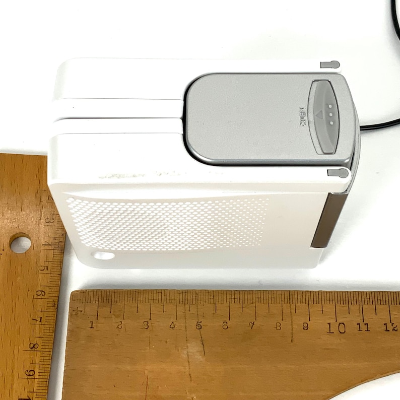 May include: A white electronic device with a gray top and a brown trim. The device has a textured pattern on the front and a black cord. A wooden ruler is placed next to the device.