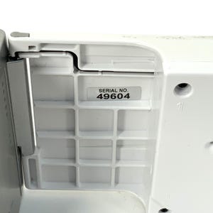 May include: Close-up of a white plastic component with a grid-like pattern. A sticker with the text "SERIAL NO. 49604" is affixed to the surface. The component has a curved edge and a metal piece.